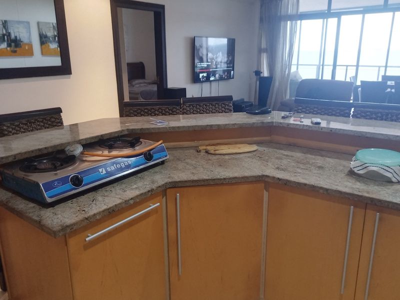 Open plan kitchen area featuring a granite breakfast bar, wooden cabinetry, and a built-in gas cooktop, overlooking the living space and balcony.