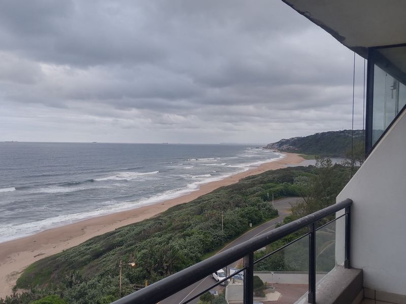 A balcony view overlooking the coastline, featuring a metal and glass balustrade with expansive ocean and beach vistas.