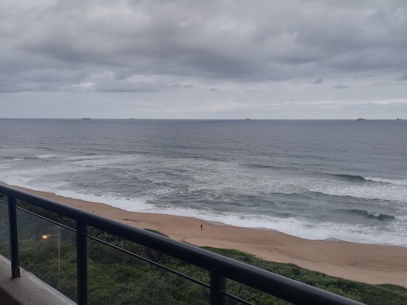 A balcony view overlooking the ocean and sandy beach, featuring a black metal railing.