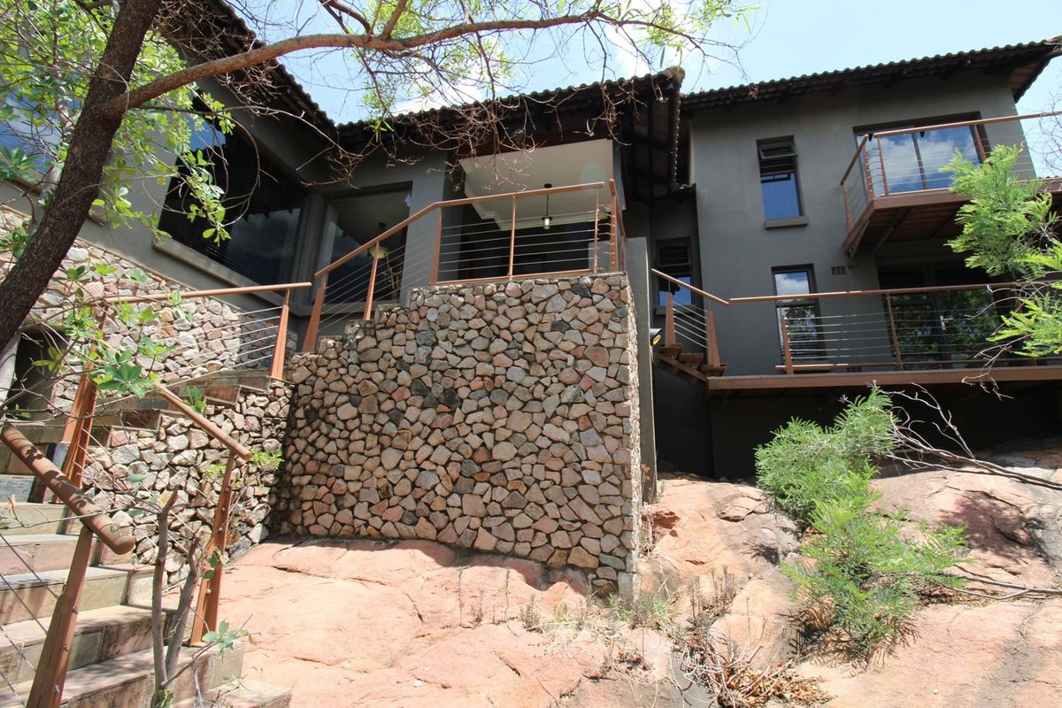 Rear exterior view of the house featuring stone retaining walls, elevated wooden decking, metal railings, and large windows set against a natural rocky landscape.
