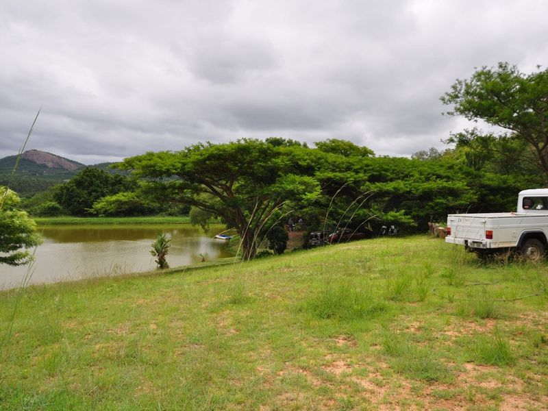 A scenic view of a farm dam surrounded by mature trees and natural grassland under a cloudy sky.