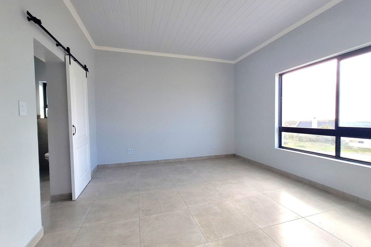 A bright, empty bedroom featuring neutral tiled flooring, a white tongue-and-groove ceiling, and a sliding barn-style door leading to an en-suite bathroom.