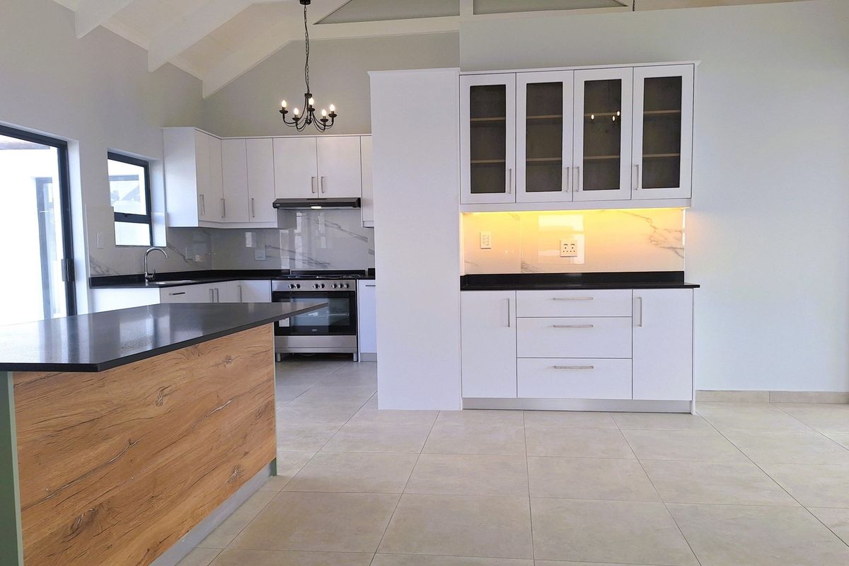 Modern open-plan kitchen featuring white cabinetry, dark stone countertops, marble-look splashbacks, integrated lighting, and a central island with wood-grain finish.