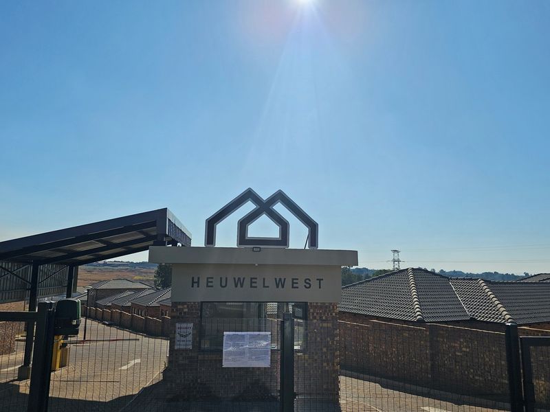 The entrance gate and security guardhouse of the Heuwelwest residential complex, featuring brick construction and a modern metal roof structure.