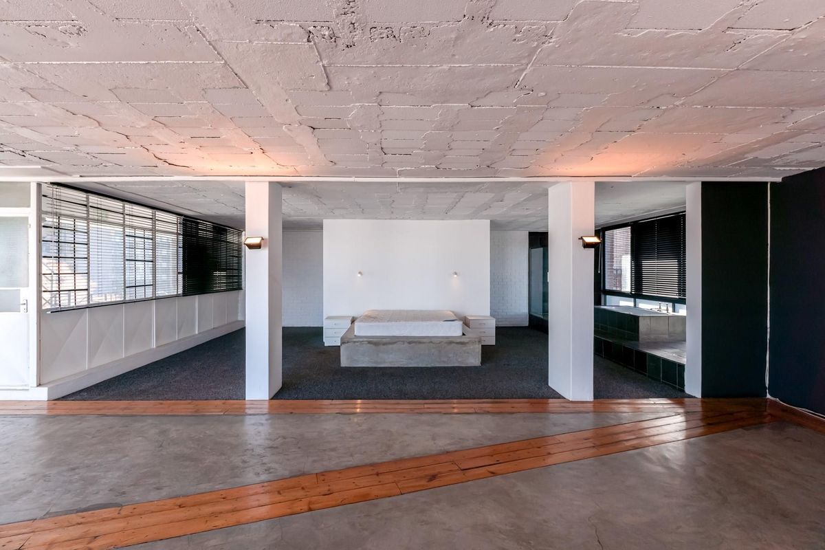 Spacious open-plan bedroom featuring a central concrete platform bed, exposed structural ceiling, and integrated bathroom area with large windows and blinds.