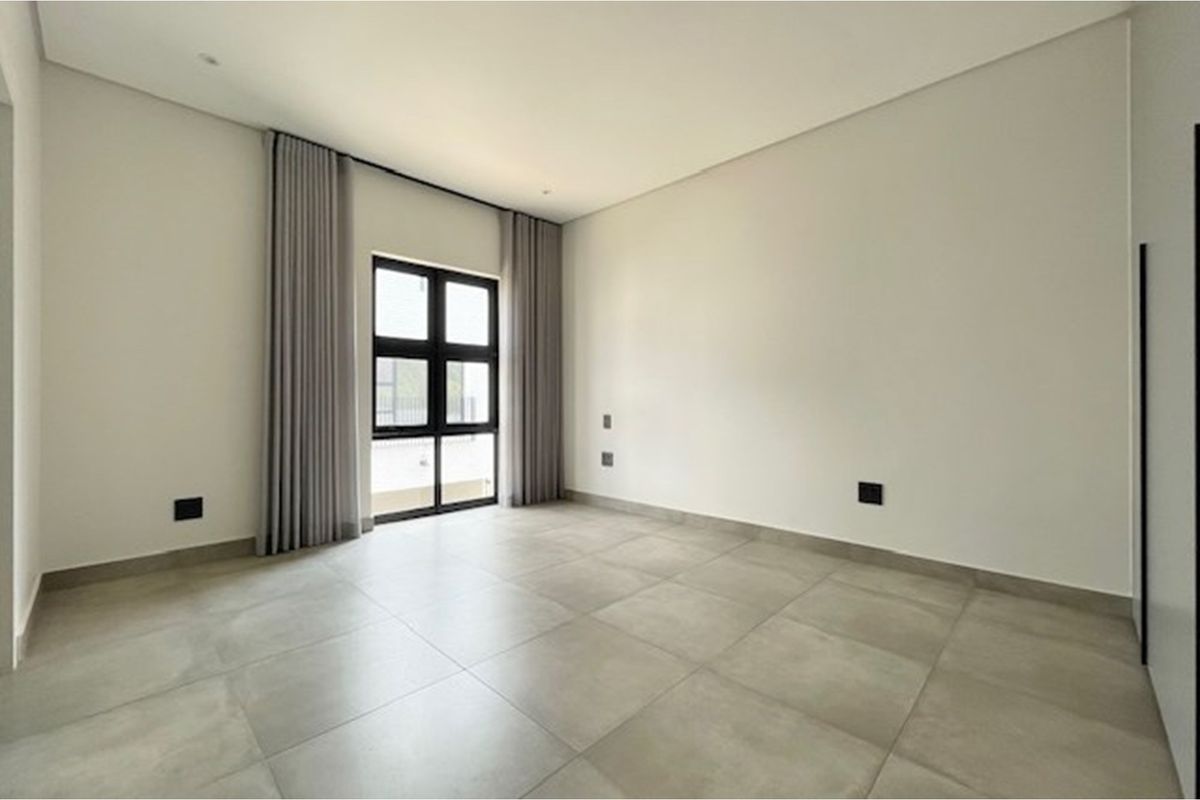 A bright, modern bedroom featuring large format floor tiles, neutral walls, and floor-to-ceiling curtains framing a window with exterior access.