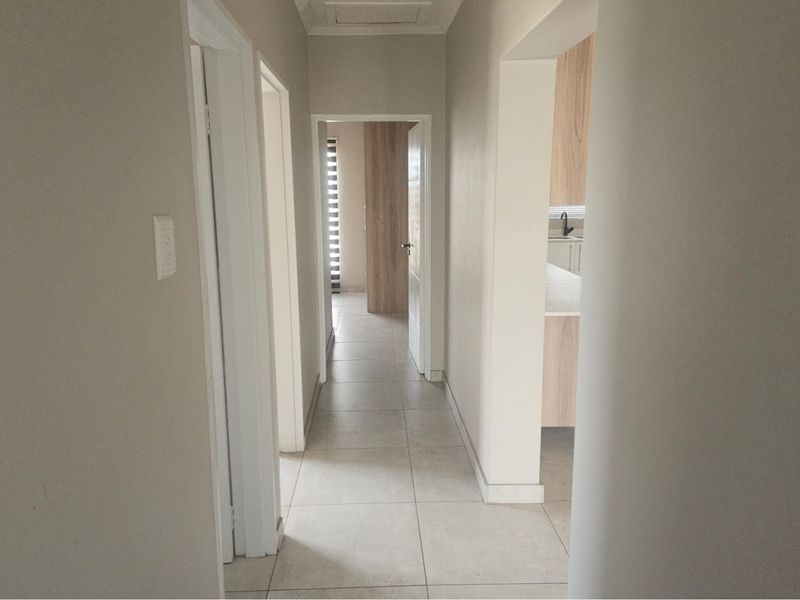 A clean, neutral-toned hallway featuring tiled flooring and access to adjacent rooms, including a glimpse into a modern kitchen area.