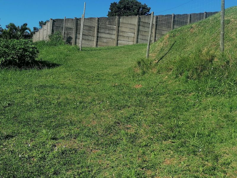 A view of a sloped residential plot featuring a grass lawn and a precast concrete boundary wall with wire fencing.