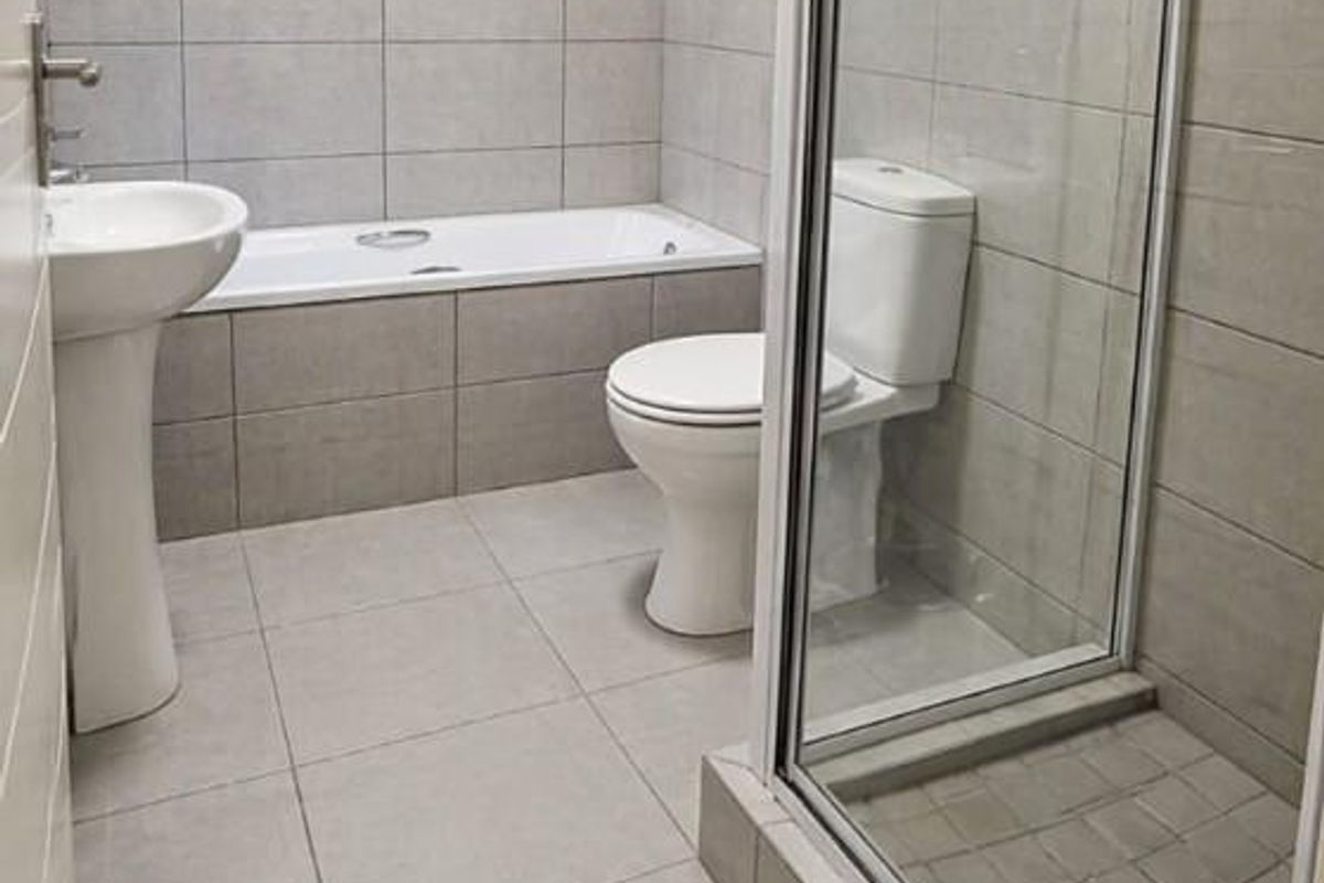A modern bathroom featuring a tiled bathtub, a separate glass-enclosed shower, a pedestal basin, and a toilet with neutral grey wall and floor tiles.