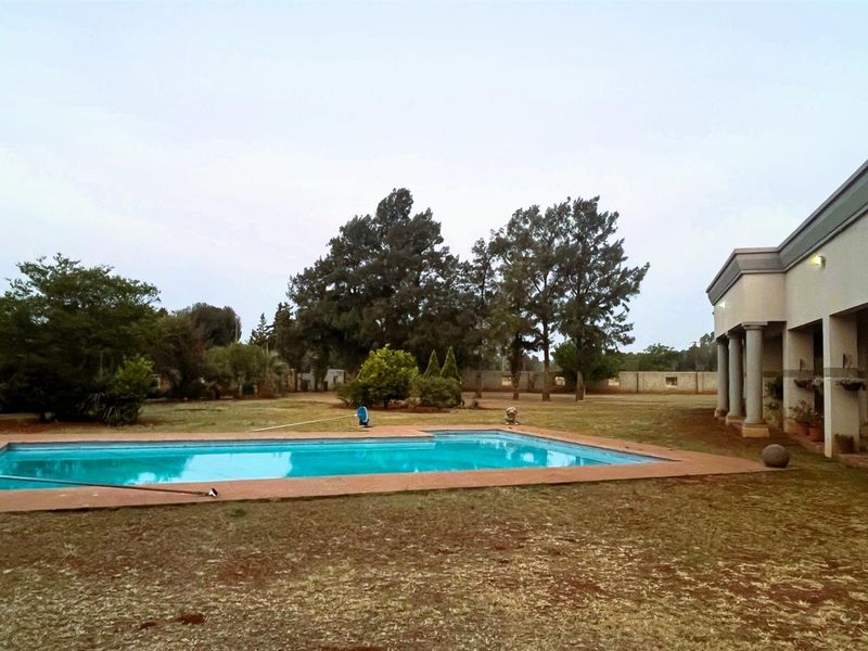 A spacious backyard featuring a rectangular swimming pool, a covered patio area with columns, and established trees on a large plot.