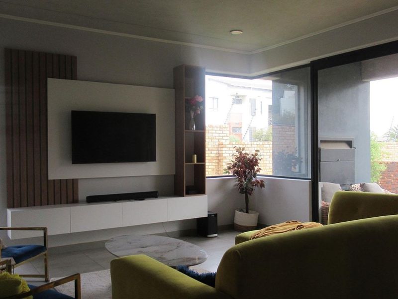 Modern living room featuring a built-in media wall with timber slat detailing, sleek cabinetry, and large corner windows providing natural light and garden views.