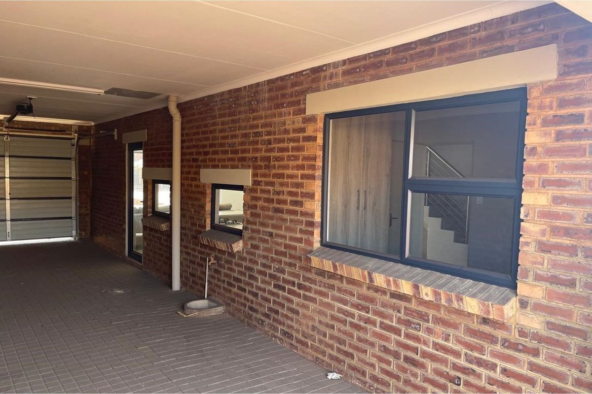 A covered exterior walkway featuring exposed brickwork, paved flooring, and access to a garage, with views into the interior through modern framed windows.