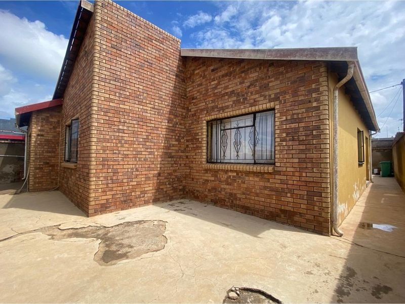 Exterior view of the property showing brick facade, security bars on windows, and concrete paved area requiring maintenance.