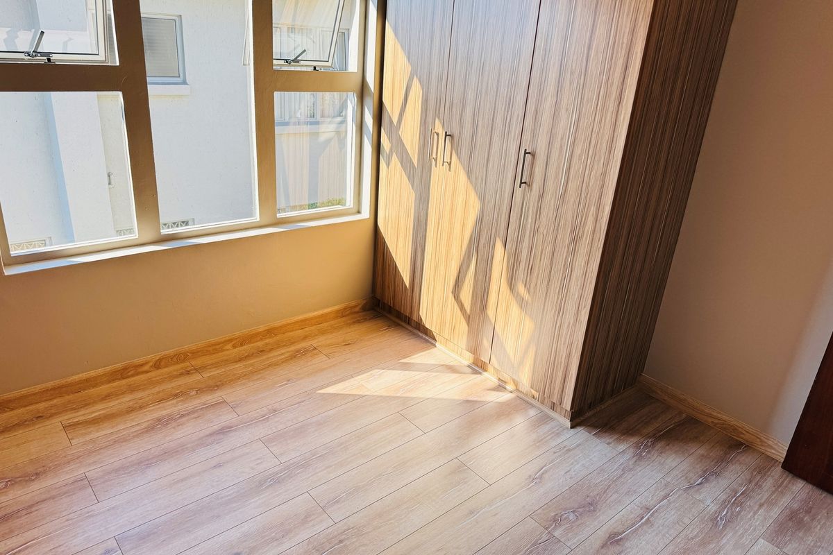 A bright, empty bedroom featuring modern laminate flooring, built-in wooden cupboards, and large aluminium-framed windows allowing for ample natural light.