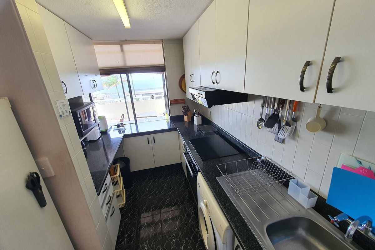 Compact galley-style kitchen featuring white cabinetry, dark granite countertops, integrated appliances, and a tiled backsplash with a view towards the exterior.