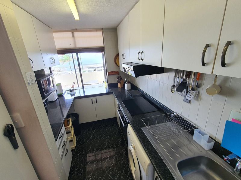 Compact galley-style kitchen featuring white cabinetry, dark granite countertops, integrated appliances, and a tiled backsplash with a view towards the exterior.