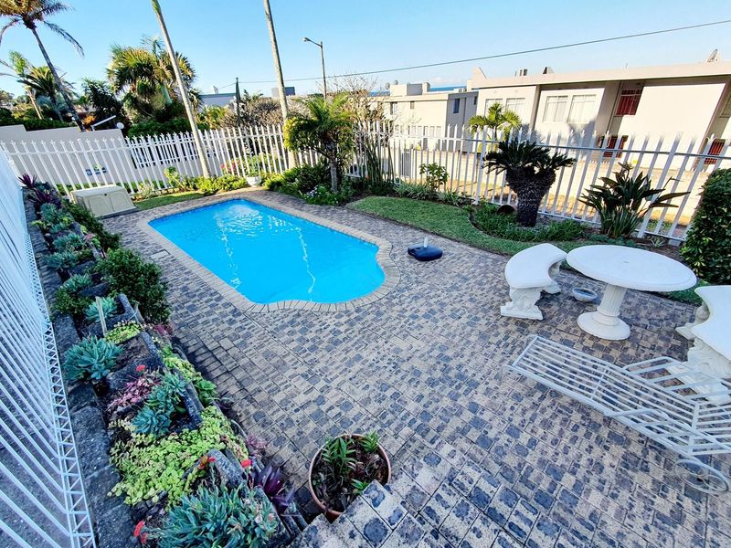 A well-maintained outdoor pool area featuring a paved patio, landscaped garden beds, and secure perimeter fencing.