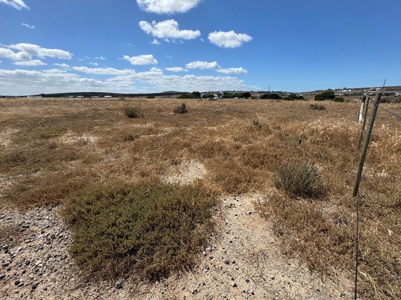 A wide-angle view of a vacant residential plot featuring dry natural vegetation and perimeter wire fencing under a clear blue sky.