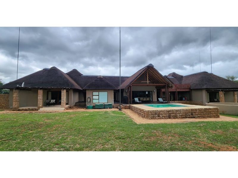 Rear view of a spacious house featuring a thatched roof, stone-clad swimming pool, and covered patio area set within a large garden.