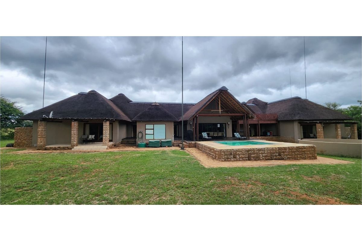 Rear view of a spacious house featuring a thatched roof, stone-clad swimming pool, and covered patio area set within a large garden.