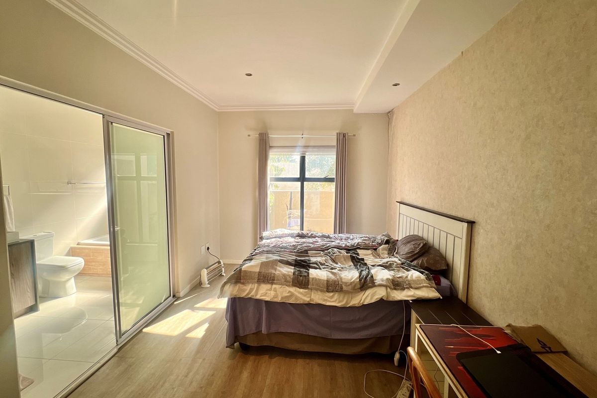 A well-lit bedroom featuring laminate flooring, a sliding glass door leading to an en-suite bathroom, and a large window providing natural light.