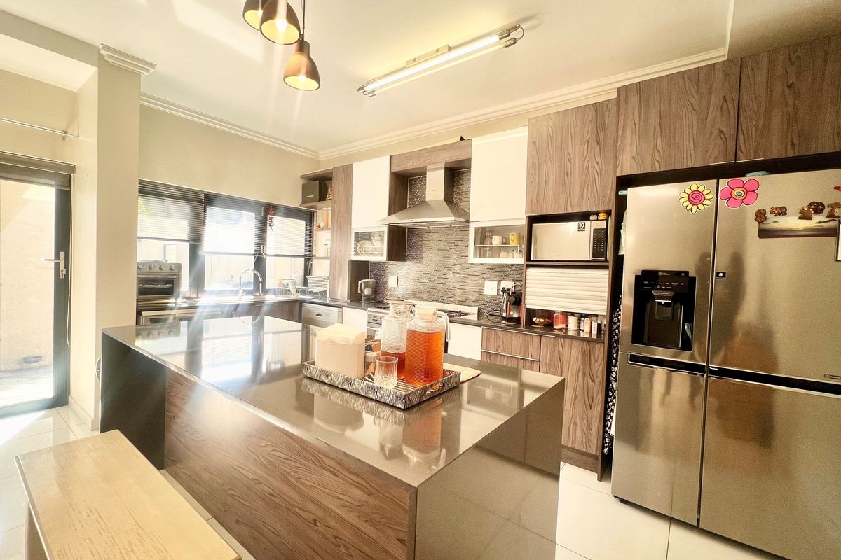 Modern kitchen featuring a large central island, wood-grain cabinetry, stone countertops, stainless steel extractor fan, and tiled flooring with direct patio access.