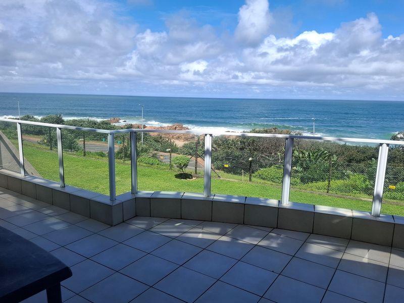 A spacious balcony featuring tiled flooring and a modern glass balustrade, offering expansive, unobstructed views of the ocean and coastline.