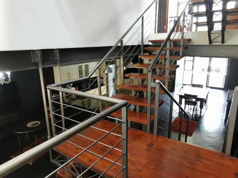 An open-plan living area featuring a prominent timber staircase with steel balustrades, exposed structural beams, and a modern kitchen layout.