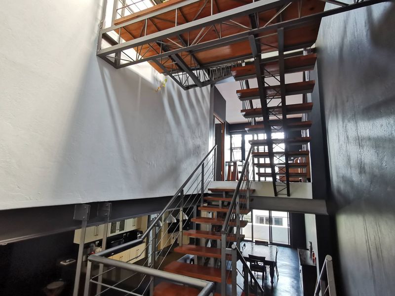 A modern industrial-style living area featuring a striking open-tread timber and steel staircase connecting to a mezzanine level with exposed structural beams.