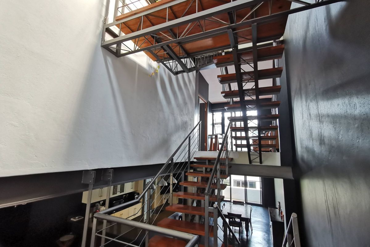 A modern industrial-style living area featuring a striking open-tread timber and steel staircase connecting to a mezzanine level with exposed structural beams.