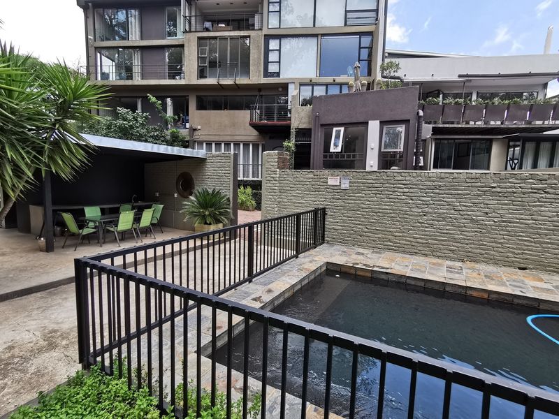 Outdoor pool area featuring a swimming pool with stone coping, metal safety fencing, and a covered patio space set against a brick boundary wall.