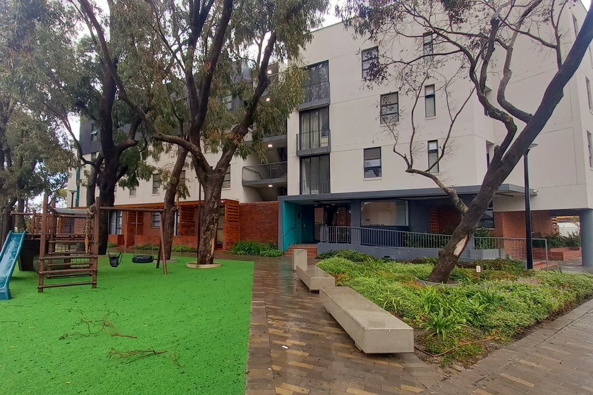 Exterior view of an apartment complex featuring a landscaped garden, paved walkways, concrete seating, and a children's play area.