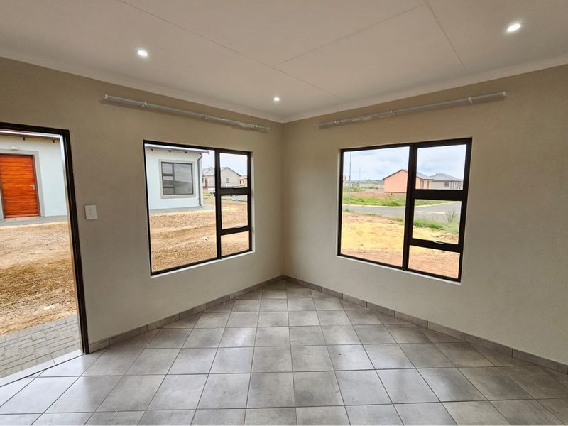 A bright, empty living area featuring neutral tiled flooring, modern aluminium-framed windows, and recessed ceiling downlights, offering a clean and versatile space.