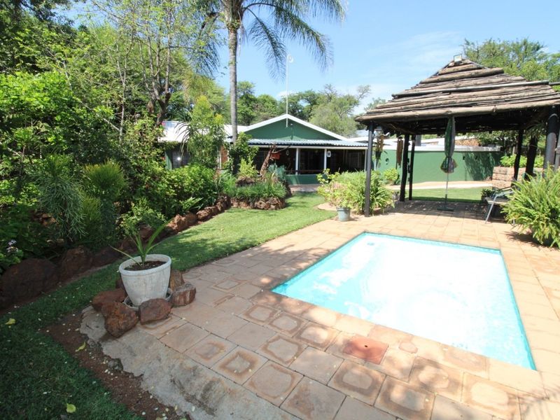 A tranquil farm garden featuring a swimming pool, paved surround, and a rustic thatched gazebo, set against a backdrop of lush greenery.
