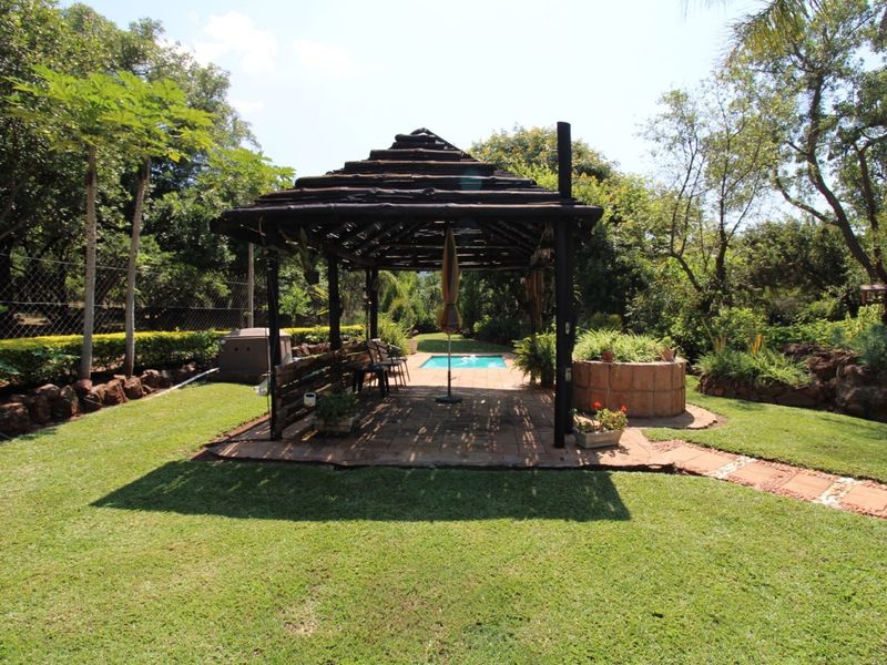 A rustic outdoor entertainment area featuring a thatched gazebo, paved patio, and a swimming pool set within a landscaped garden.
