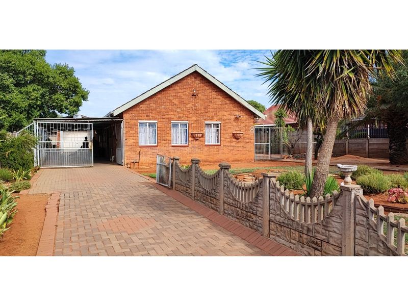 Front view of a single-story brick house featuring a paved driveway, secure metal gate, and established garden with precast boundary walling.