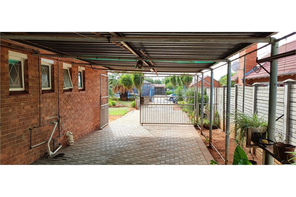 A covered carport area featuring brick walling, paved flooring, and a metal security gate leading to the rear garden space.