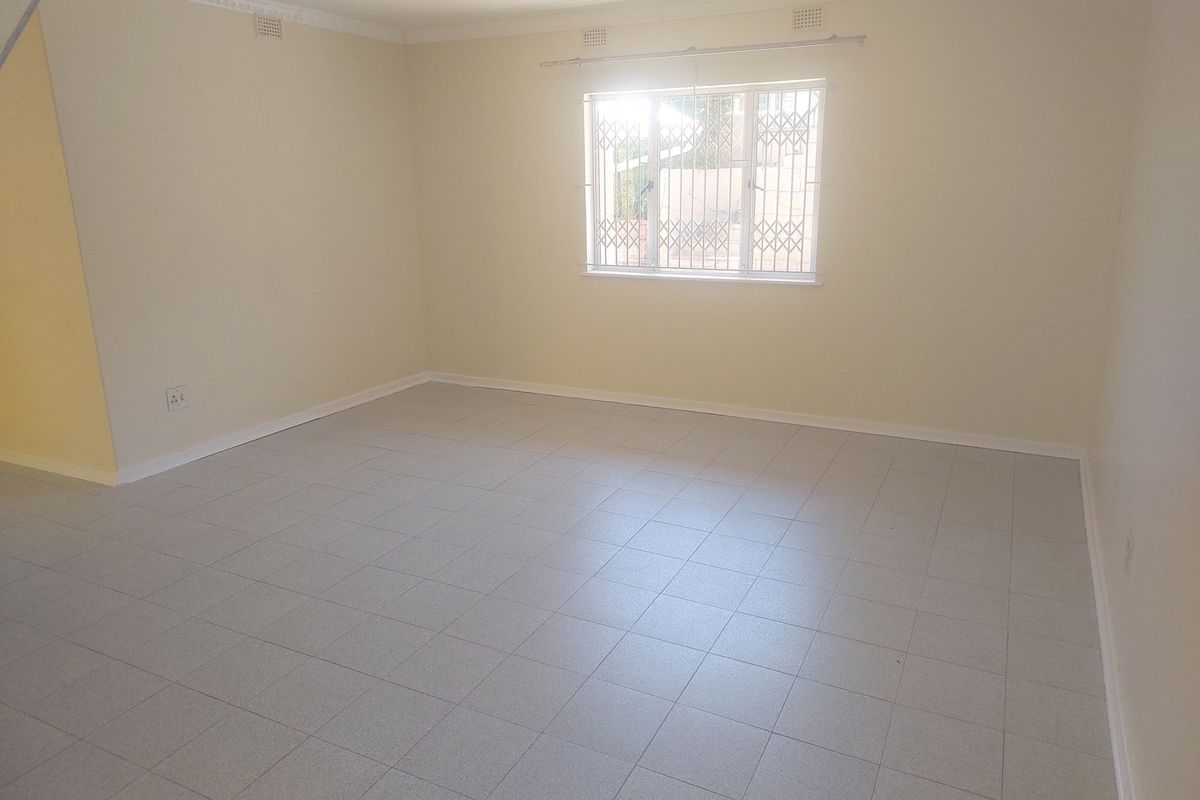 A spacious, empty living area featuring neutral tiled flooring, crown molding, and a window fitted with security bars.