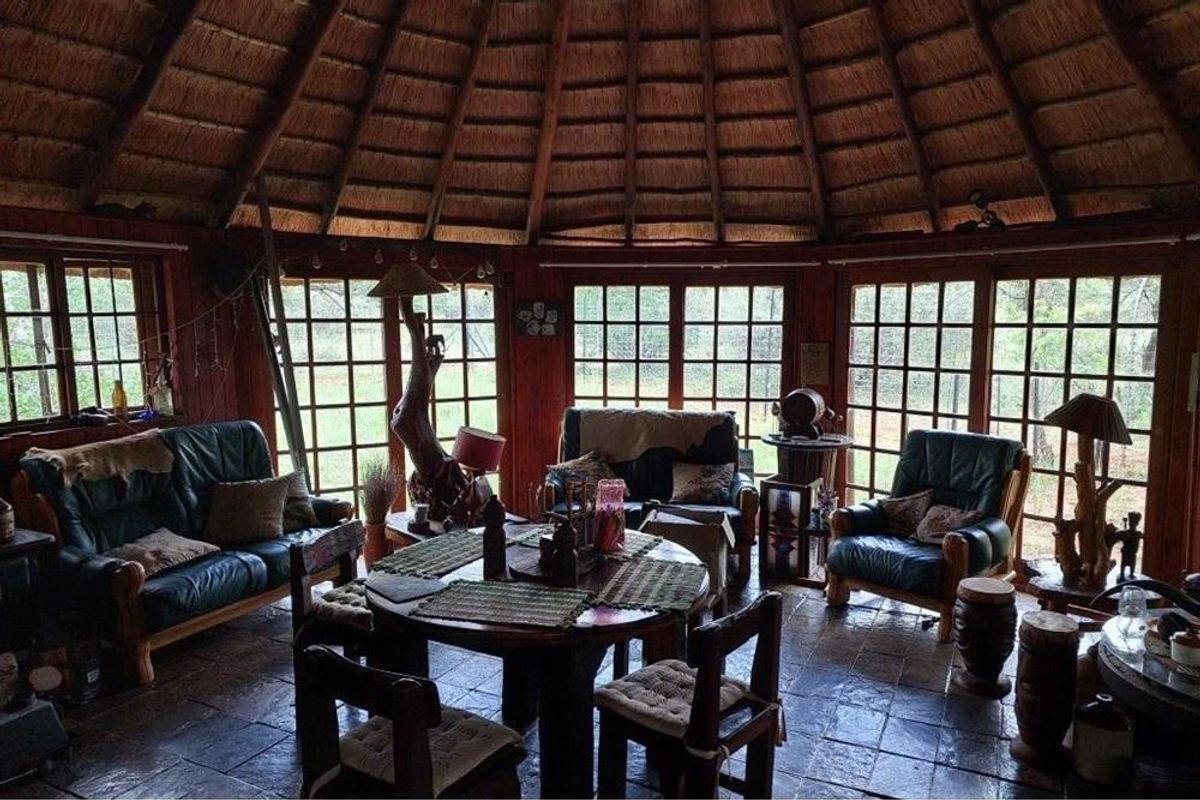 A rustic circular living area featuring a traditional thatched roof, exposed timber walls, and large grid-pane windows overlooking the surrounding farm landscape.