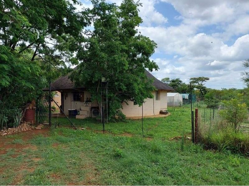 A view of a rustic thatched-roof farm dwelling set within a natural, grassy landscape with mature trees and perimeter fencing.