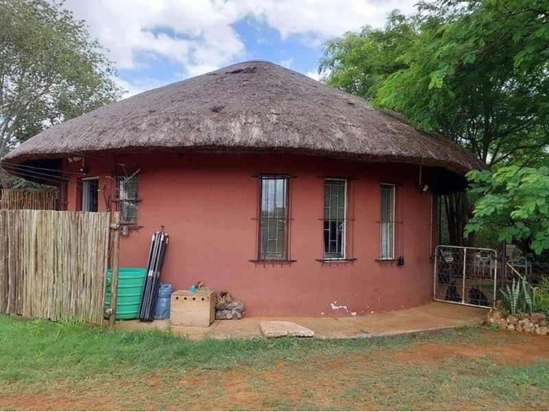 A traditional round farm cottage featuring a thatched roof, red exterior walls, and security bars on the windows, set within a natural garden.