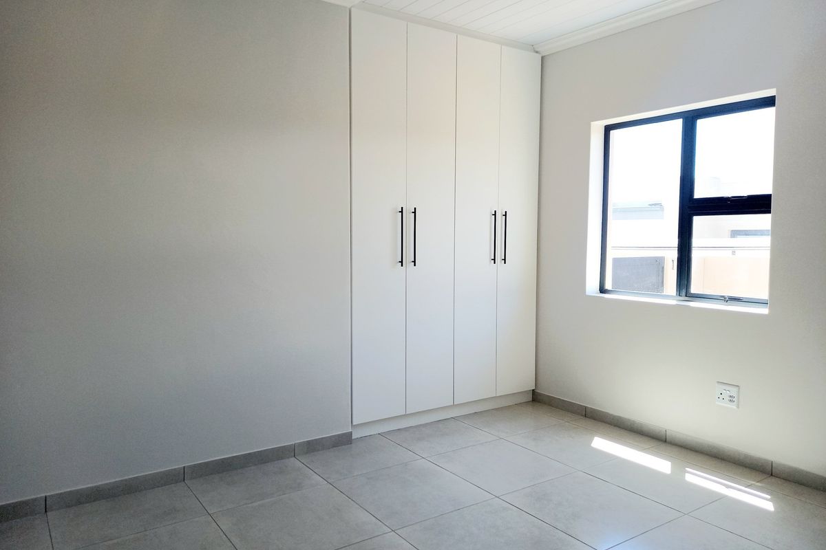 A bright, modern bedroom featuring neutral tiled flooring, clean white walls, and ample built-in cupboard storage with contemporary black handles.