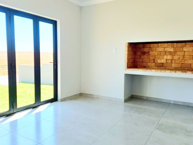 A bright, empty covered patio featuring tiled flooring, a built-in brick braai, and large aluminium sliding doors leading to the garden.