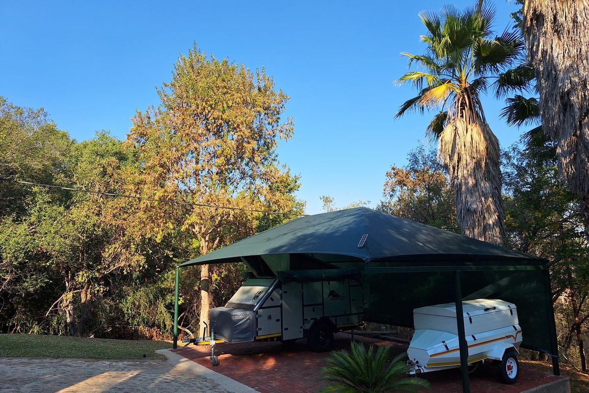 Outdoor shaded parking area featuring a permanent steel-framed shade cloth structure, suitable for vehicle or trailer storage within a landscaped garden setting.