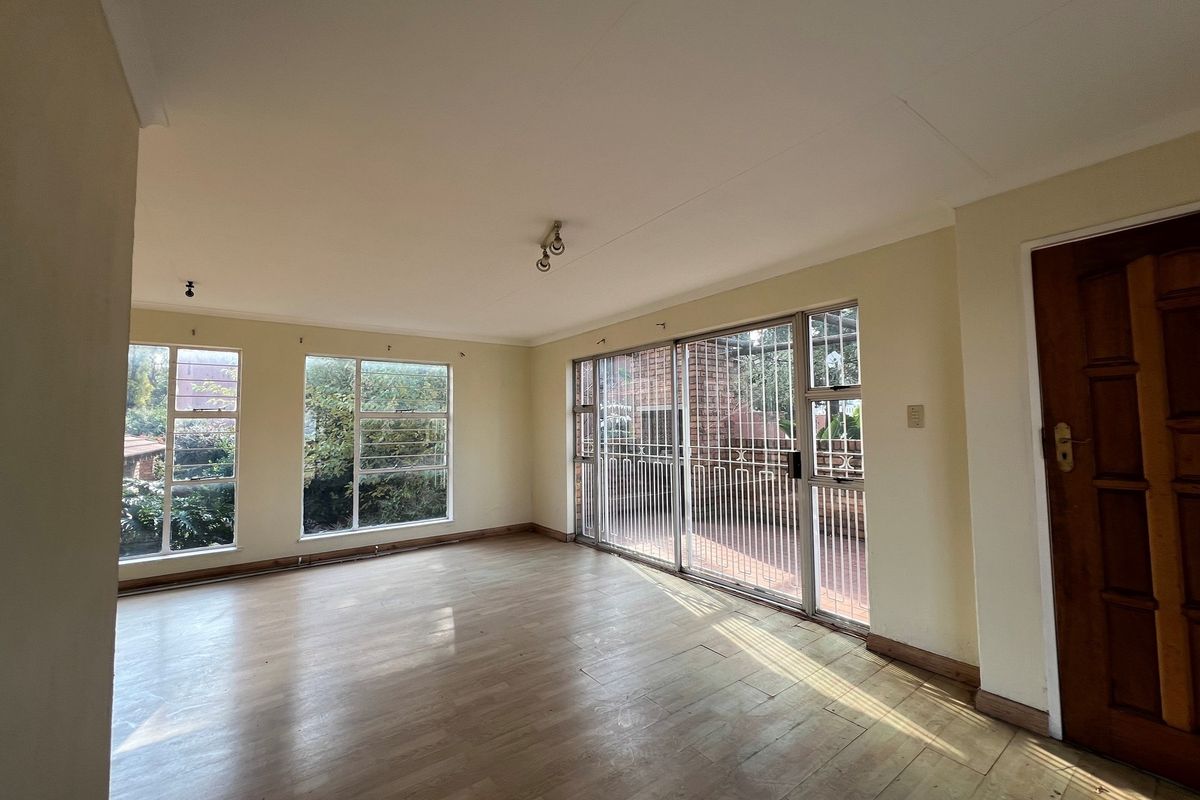 A spacious, sunlit living room featuring light-toned laminate flooring, large windows, and a sliding glass door providing access to an outdoor patio area.