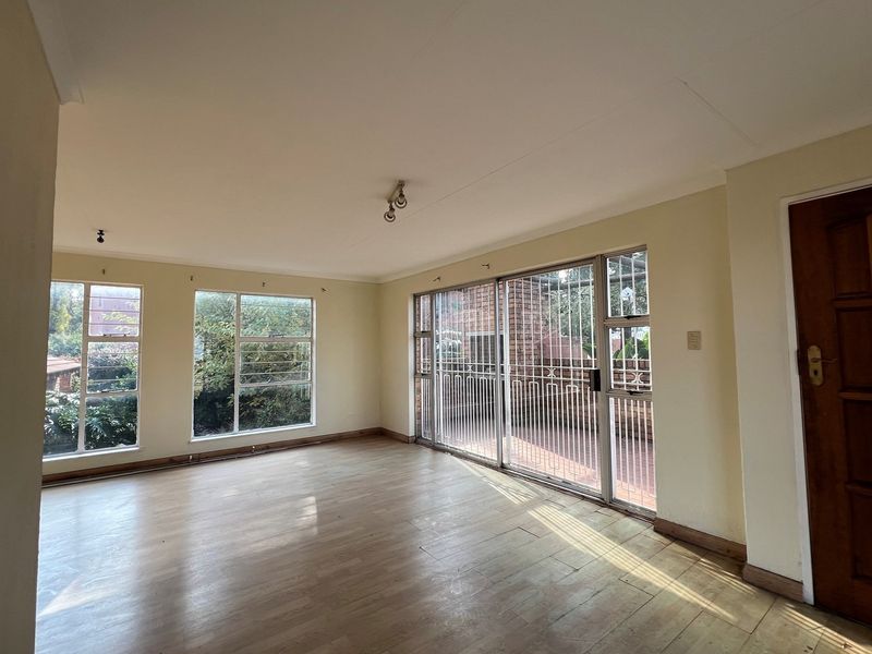 A spacious, sunlit living room featuring light-toned laminate flooring, large windows, and a sliding glass door providing access to an outdoor patio area.