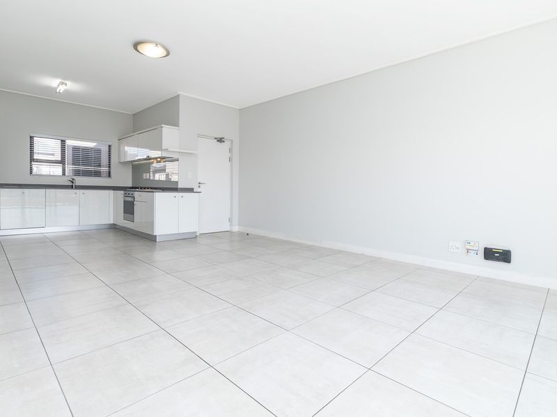 A bright, open-plan living and kitchen area featuring modern tiled flooring, sleek white cabinetry, and integrated kitchen appliances in a clean, vacant space.