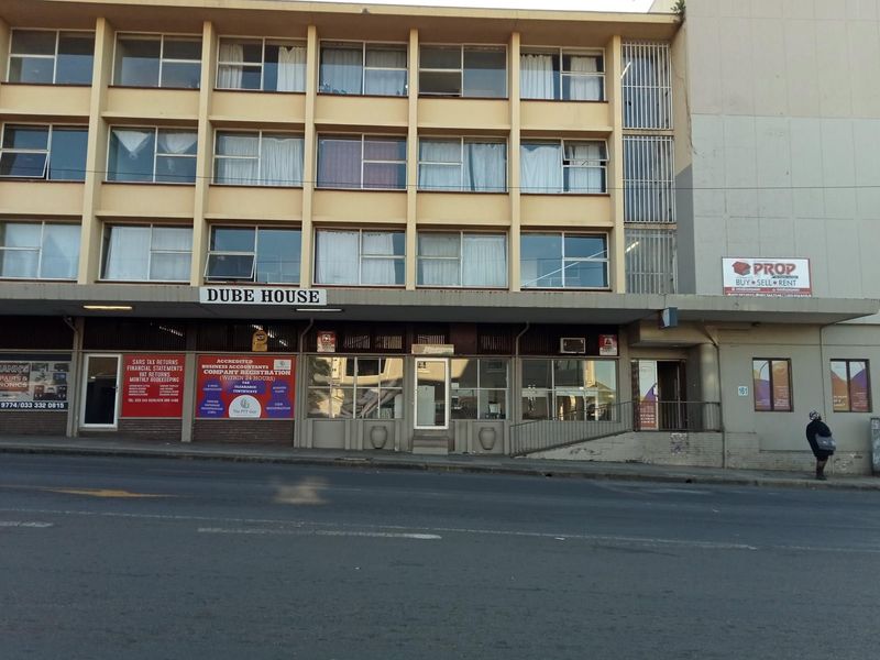 Street-level view of Dube House, a multi-story apartment and commercial building featuring a concrete facade and ground-floor retail spaces.
