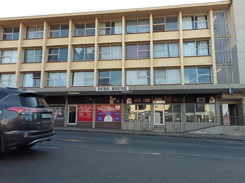 Street-level view of Dube House, a multi-story apartment building featuring ground-floor commercial units and a concrete facade.