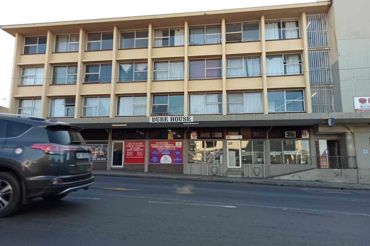 Street-level view of Dube House, a multi-story apartment building featuring ground-floor commercial units and a concrete facade.